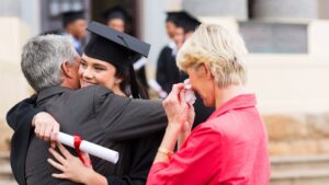 Young woman, wearing graduation attire, hugging her father with her mother shedding tears of happiness.